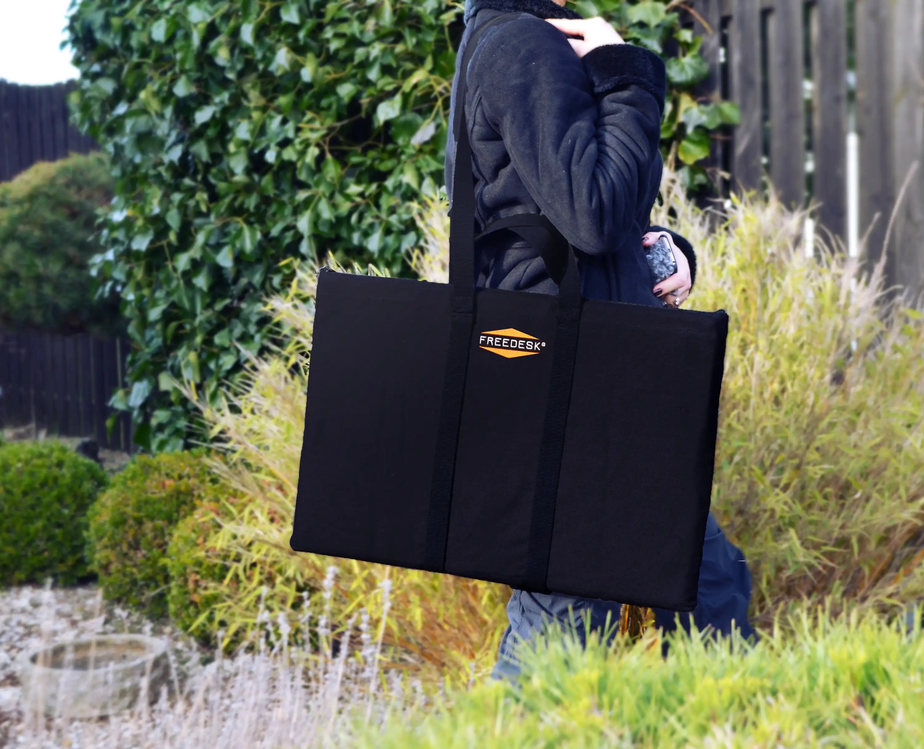 Freedesk Carrying Case Compact in black, designed for convenient transport of the Freedesk Desk Riser, shown being carried outdoors.